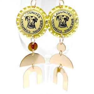 May include: A pair of dangle earrings featuring repurposed beer bottle caps with a dog logo. The earrings have gold-colored metal accents, including a small circle, a half-circle, and a U-shaped design. The bottle caps are yellow with black text.