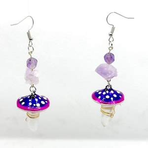 May include: A pair of dangle earrings featuring a purple and pink mushroom charm with white polka dots, a clear crystal, and amethyst stones.