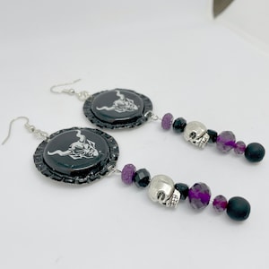 May include: A pair of black and silver earrings with a skull charm and purple beads. The earrings are made from bottle caps with a white image of a horned creature.