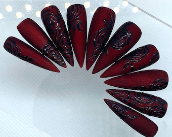 May include: A set of 10 burgundy-colored false nails with black rose designs. The nails are long and pointed.