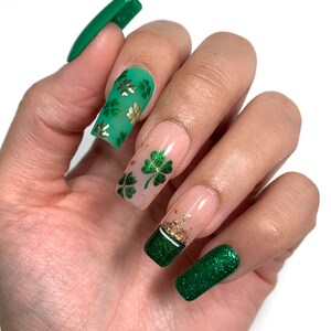 Puede incluir: Primer plano de una mano con uñas largas, verdes y nude decoradas con diseños de trébol de cuatro hojas dorados. Algunas uñas tienen brillo.