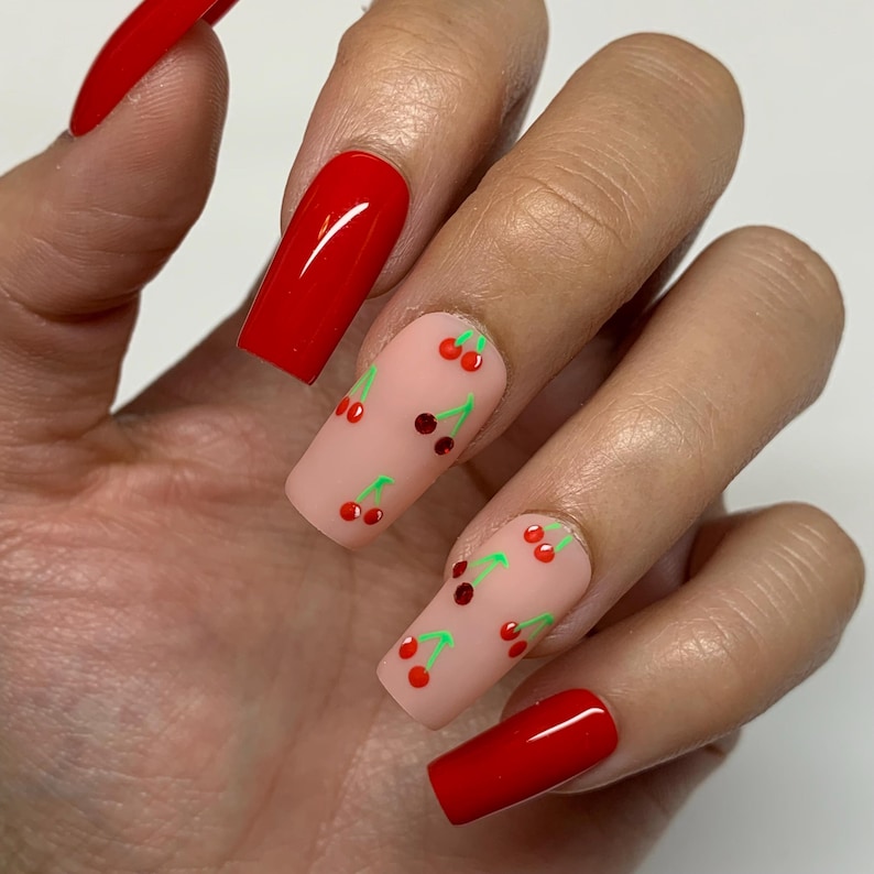 May include: Close-up of a hand with long, square-shaped nails. Some nails are painted red, while others are painted a light pink with a cherry design.