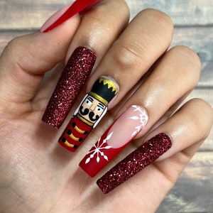 May include: A hand with long, square-shaped, painted nails. The nails feature a Christmas theme with a nutcracker design, red glitter, and snowflake accents. The nails are painted in red, white, black, and gold.