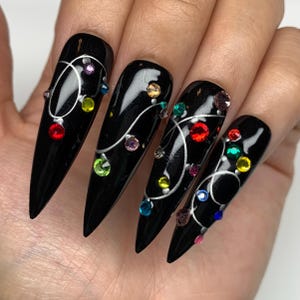 Peut inclure: Ongles noirs en forme de stiletto avec une finition brillante, ornés d'un motif tourbillonnant argenté et de strass colorés. Les strass sont de différentes couleurs, dont le rouge, le jaune, le vert, le bleu et le rose, ajoutant de l'éclat.