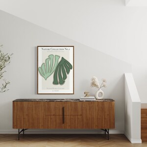Könnte beinhalten: Ein gerahmtes Poster mit einem minimalistischen Design, das zwei grüne Blätter auf einem cremefarbenen Hintergrund zeigt. Das Poster trägt den Titel "Nature Collection No.1" und hängt an einer weißen Wand über einem Holzschrank mit schwarzen Metallbeinen. Im Vordergrund steht eine kleine Topfpflanze.