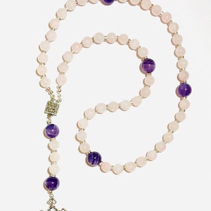May include: A rosary featuring light pink rose quartz beads and larger purple amethyst beads. The rosary has a silver-coloured metal cross and connecting pieces. The beads are strung on a silver-coloured chain.
