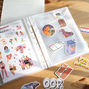 May include: A clear plastic binder with a white background and a variety of colorful stickers. The stickers feature illustrations of people, flowers, and other designs. The binder has a ring mechanism and is open to reveal the stickers inside.