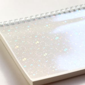 Könnte beinhalten: Ein spiralgebundenes Notizbuch mit weißem Einband und schillernden Glitzersternen. Die Seiten des Notizbuchs sind weiß, und die Spiralbindung ist silberfarben. Der Einband hat eine transparente Schutzauflage.