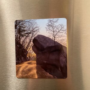 May include: A photograph of a large, dark rock formation in a forest setting. The rock is partially obscured by trees and branches, and the sky is a soft blue with hints of pink and orange. The ground is covered in fallen leaves and the light is soft and warm.