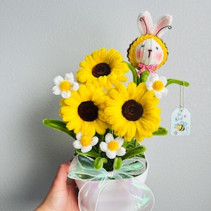 Può includere: Un vaso bianco contenente un bouquet di girasoli gialli e margherite bianche realizzati con scovolini. Un piccolo coniglio di peluche vestito da ape si trova tra i fiori. Un'etichetta bianca pende con la scritta Be Kind e un'ape disegnata.