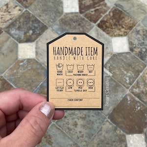 May include: Brown paper tag with black text and icons for handmade item care instructions. The tag says "Handmade Item Handle With Care" and includes icons for hand wash, cold wash, warm wash, hot wash, lay flat to dry, low tumble dry, medium tumble dry, and high tumble dry. The tag also says "Fiber Content" at the bottom.