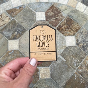 Könnte beinhalten: Ein braunes Papieretikett mit schwarzem Text, der "Fingerlose Handschuhe 100% handgefertigt Größe: Kind - Teen - Erwachsener" lautet und eine kleine Illustration von zwei Fingerlosen Handschuhen zeigt. Das Etikett ist in der unteren rechten Ecke mit "Smart Creative Living" beschriftet.