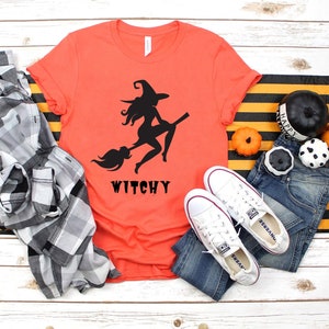 May include: A coral orange t-shirt with a black silhouette of a witch riding a broomstick. The text "Witchy" is printed below the image.