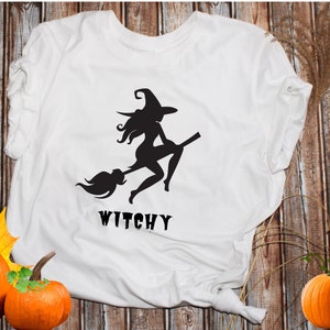 May include: White t-shirt featuring a black silhouette of a witch riding a broomstick. The text "Witchy" is printed beneath the image.