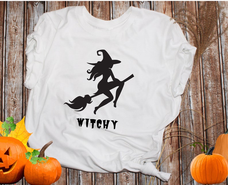 May include: White t-shirt featuring a black silhouette of a witch riding a broomstick. The text "Witchy" is printed beneath the image.