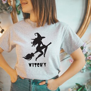 May include: A grey t-shirt featuring a black silhouette of a witch flying on a broomstick. The text "Witchy" is printed below the image.