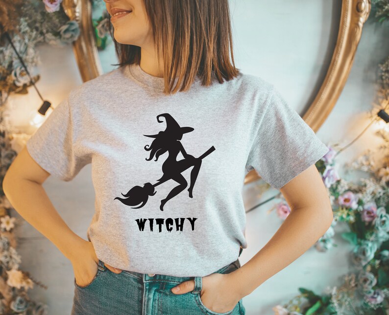 May include: A grey t-shirt featuring a black silhouette of a witch flying on a broomstick. The text "Witchy" is printed below the image.