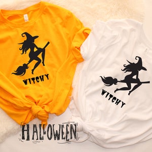May include: Two t-shirts featuring a witch silhouette design with the word "Witchy" printed on them. The shirts are yellow and white. The word "Halloween" is written in black on a white background.