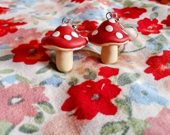 May include: A pair of red and white mushroom earrings with silver hooks. The mushrooms have white dots on the red caps and a cream-colored stem.