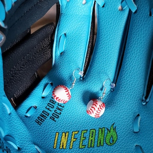 May include: A pair of baseball-themed earrings with red stitching on a white background. The earrings are hanging from a blue baseball glove with the word "INFERNO" printed on it.