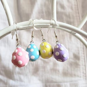 May include: Four pairs of dangle earrings with pastel colored egg charms. The eggs are decorated with white polka dots. The colors of the eggs are pink, blue, yellow, and purple.