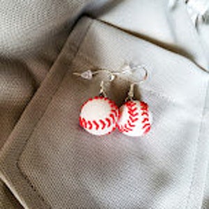 May include: A pair of silver baseball earrings with white and red baseballs. The earrings are hanging from a white fabric background.
