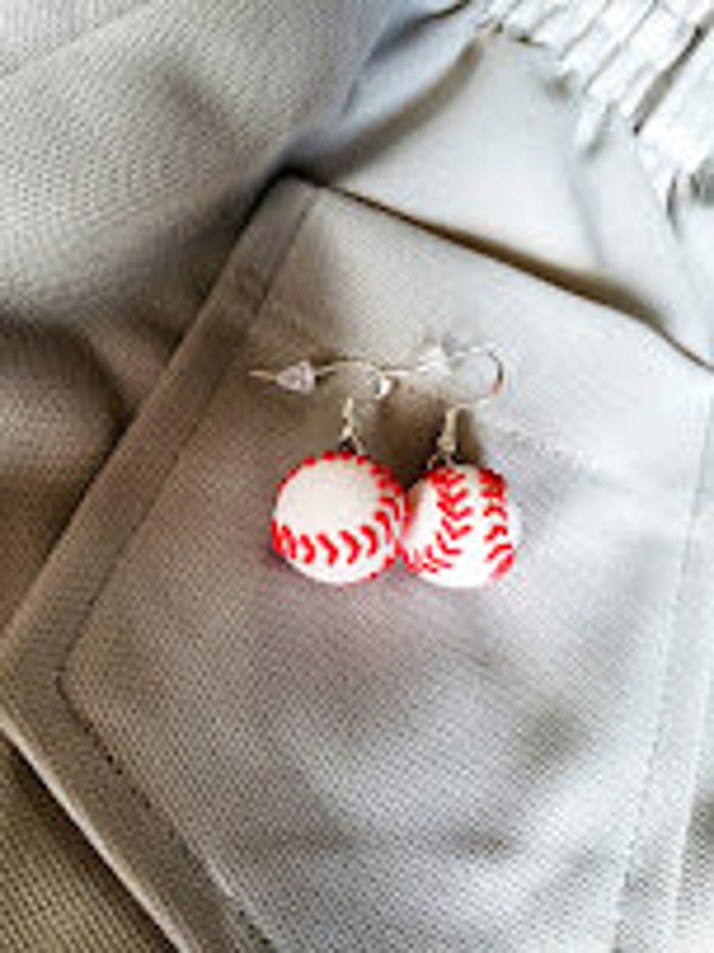 May include: A pair of silver baseball earrings with white and red baseballs. The earrings are hanging from a white fabric background.