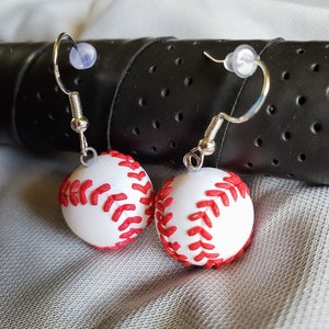 May include: A pair of silver dangle earrings with white and red baseball charms. The baseball charms have red stitching.