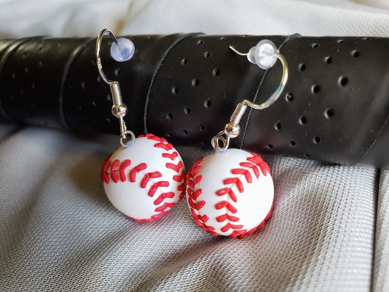 May include: A pair of silver dangle earrings with white and red baseball charms. The baseball charms have red stitching.