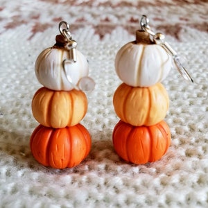 May include: A pair of dangle earrings featuring three stacked polymer clay pumpkins. The pumpkins are white, orange, and a darker orange. The earrings are made with silver hooks.