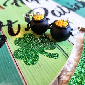 May include: A pair of gold pot of gold earrings with a shamrock on a green and brown background. The earrings are made of black polymer clay and have gold glitter inside.