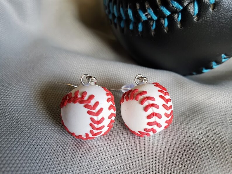 May include: A pair of silver earrings with white baseball charms. The baseball charms have red stitching.