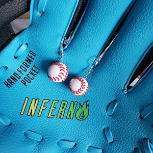 May include: A blue baseball glove with white stitching and the words "Hand Formed Pocket" and "Inferno" printed on it. Two white baseball earrings with red stitching are hanging from the glove.