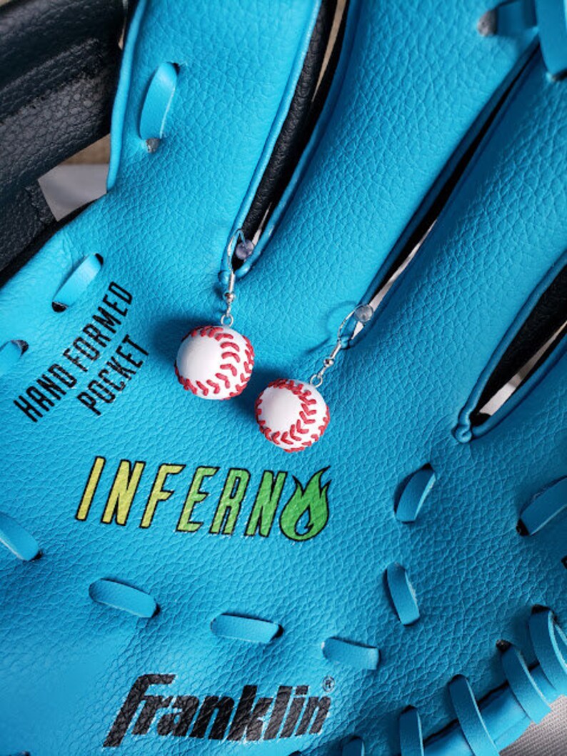 May include: A blue baseball glove with white stitching and the words "Hand Formed Pocket" and "Inferno" printed on it. Two white baseball earrings with red stitching are hanging from the glove.