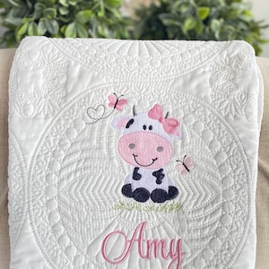 May include: White baby blanket with a quilted design. Features an embroidered cartoon cow with a pink bow and butterflies. The name "Amy" is embroidered in pink script below the cow.
