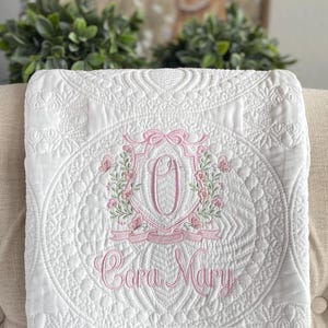 May include: White quilted blanket with a pink floral crest and the name "Cora Mary" embroidered in pink script. The blanket features intricate stitching patterns and a decorative border, perfect for a baby.