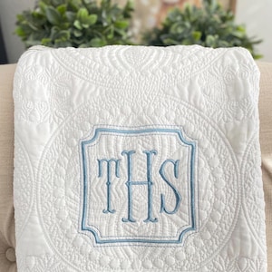 May include: White quilted blanket with a detailed pattern and a central monogram in light blue. The monogram is framed within a decorative border. The blanket is folded, showcasing the intricate stitching and design.