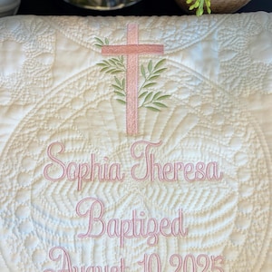 K&ouml;nnte beinhalten: Cremefarbene, gesteppte Taufdecke mit rosa gestickten Details. Mit einem Kreuz mit Olivenzweigen, dem Namen "Sophia Theresa" und dem Datum "10. August 2025". Die Decke hat ein kreisf&ouml;rmiges, florales Design.