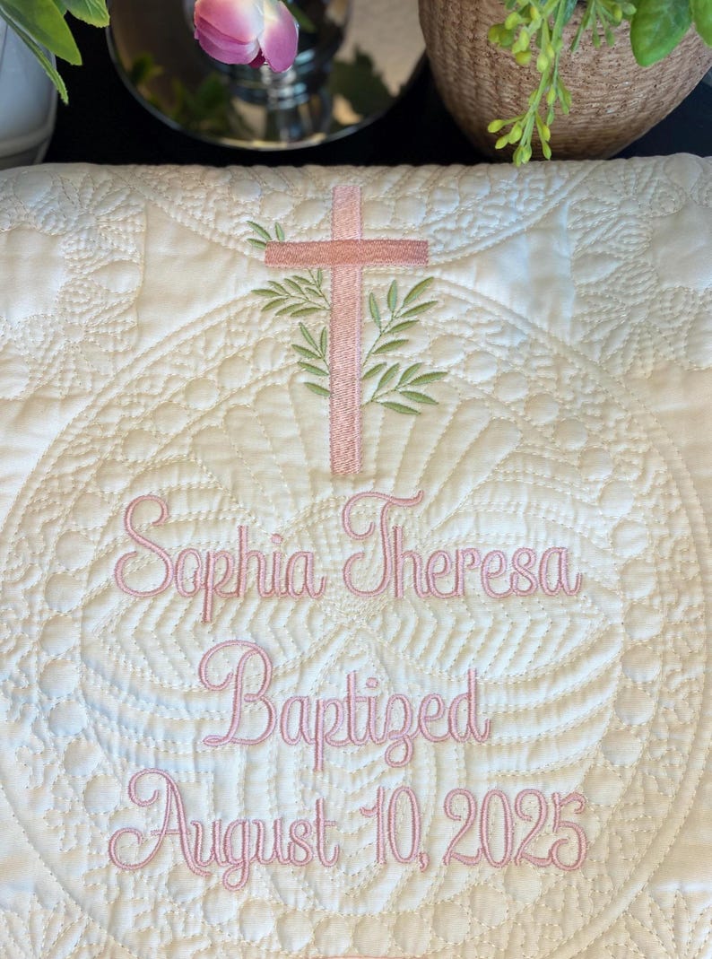K&ouml;nnte beinhalten: Cremefarbene, gesteppte Taufdecke mit rosa gestickten Details. Mit einem Kreuz mit Olivenzweigen, dem Namen "Sophia Theresa" und dem Datum "10. August 2025". Die Decke hat ein kreisf&ouml;rmiges, florales Design.