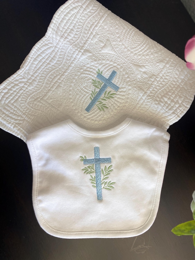 K&ouml;nnte beinhalten: Wei&szlig;es Baby-L&auml;tzchen- und Decken-Set. Das L&auml;tzchen und die Decke haben ein hellblaues Kreuzdesign mit gr&uuml;nen Bl&auml;ttern. Die Decke hat ein gestepptes Muster.