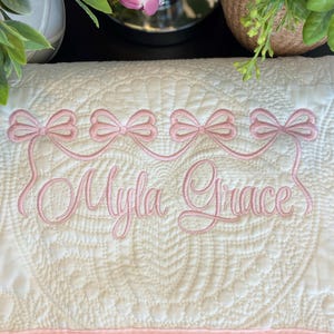 May include: Cream-colored, quilted baby blanket with pink embroidered bows and the name "Myla Grace". The blanket features a pink border and a detailed quilted pattern. A decorative item for a nursery.