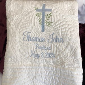 K&ouml;nnte beinhalten: Cremefarbene, gesteppte Decke mit gestickten Details. Sie zeigt ein hellblaues Kreuz mit Olivenzweigen und den Namen "Thomas John" sowie das Taufdatum "May 3, 2022" in hellblauem Faden. Die Decke hat ein dekoratives, gen&auml;htes Muster.
