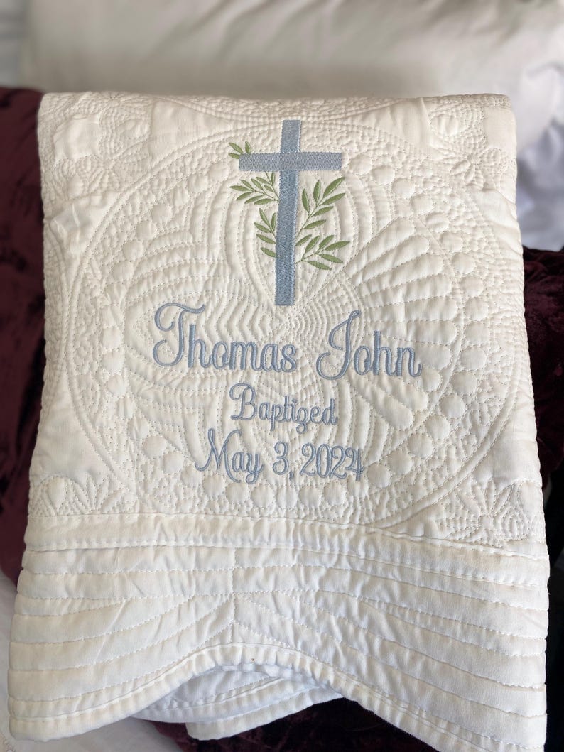 K&ouml;nnte beinhalten: Cremefarbene, gesteppte Decke mit gestickten Details. Sie zeigt ein hellblaues Kreuz mit Olivenzweigen und den Namen "Thomas John" sowie das Taufdatum "May 3, 2022" in hellblauem Faden. Die Decke hat ein dekoratives, gen&auml;htes Muster.