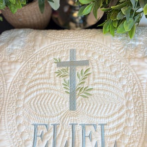 K&ouml;nnte beinhalten: Cremefarbene, gesteppte Decke mit einem detaillierten, kreisf&ouml;rmigen Muster. Ein hellblaues Kreuz mit gr&uuml;nen Bl&auml;ttern ist in der Mitte gestickt, &uuml;ber dem Namen "ELIEL" in Hellblau. F&uuml;r ein Baby oder Kind.