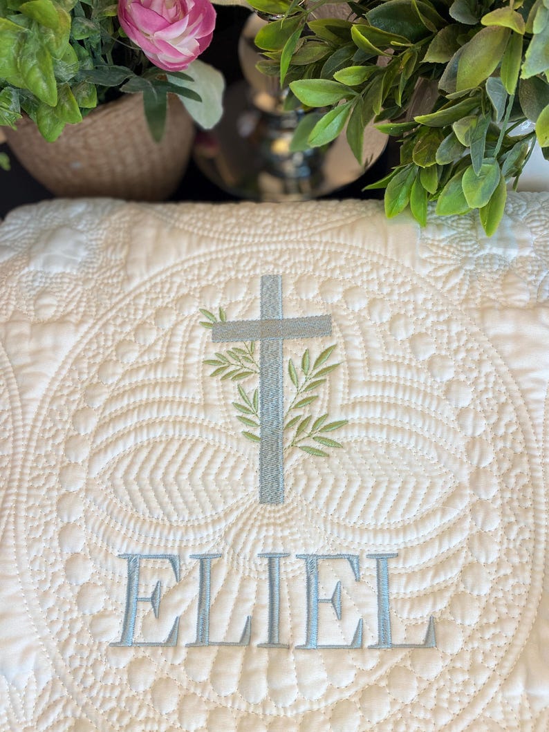 K&ouml;nnte beinhalten: Cremefarbene, gesteppte Decke mit einem detaillierten, kreisf&ouml;rmigen Muster. Ein hellblaues Kreuz mit gr&uuml;nen Bl&auml;ttern ist in der Mitte gestickt, &uuml;ber dem Namen "ELIEL" in Hellblau. F&uuml;r ein Baby oder Kind.