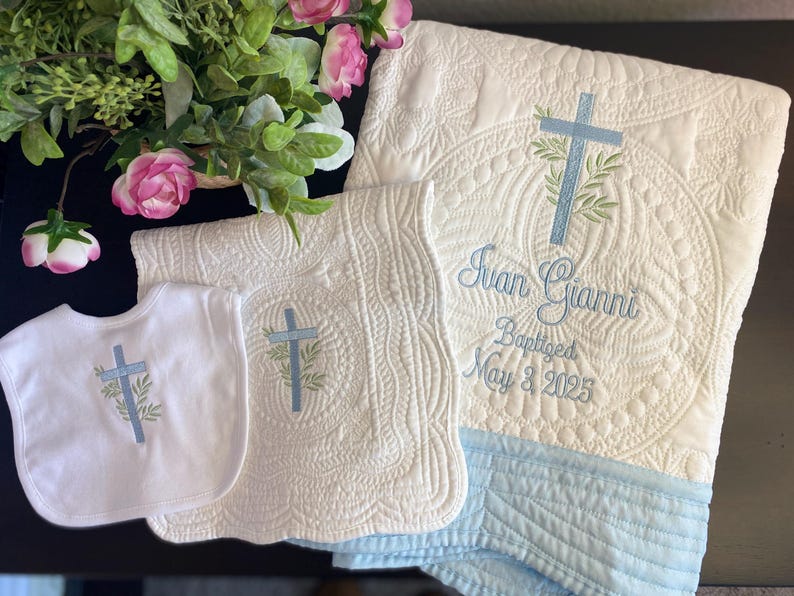 K&ouml;nnte beinhalten: Ein wei&szlig;es, gestepptes Taufset mit Decke, L&auml;tzchen und Spucktuch. Jedes Teil ist mit einem blauen Kreuz und Olivenzweig-Design bestickt. Die Decke ist mit einem Namen und Taufdatum personalisiert.