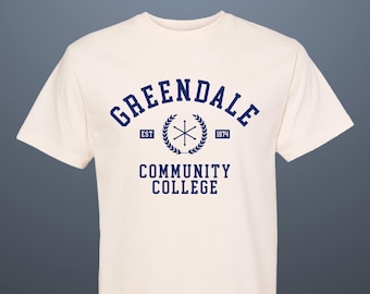 Puede incluir: Una camiseta blanca con una impresión azul marino que dice "Greendale Community College" con un escudo que presenta una estrella y una corona de laurel. El texto "Est. 1974" está impreso sobre el escudo y "Est." está impreso debajo del escudo.