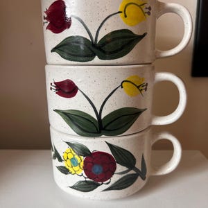 Könnte beinhalten: Drei gestapelte Keramikbecher mit gesprenkelter weißer Oberfläche. Jeder Becher ist mit handgemalten Blumendesigns in Rot, Gelb und Blau mit grünen Blättern verziert. Die Becher haben ein klassisches Griffdesign.