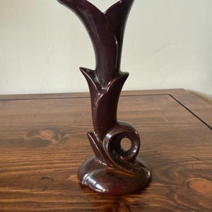 Puede incluir: Un jarrón de cerámica burdeos oscuro con una forma única y estilizada. El jarrón tiene una parte superior acampanada, un cuello delgado y un remolino decorativo en la base. El acabado brillante realza el color intenso y añade atractivo vintage.