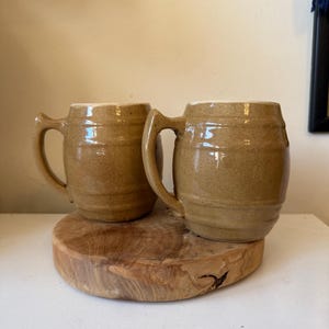 May include: Two brown ceramic beer steins with handles, sitting on a round wooden slab. The steins have a glossy finish and a classic, rustic design, perfect for serving beverages. The mugs are a warm, earthy tone.